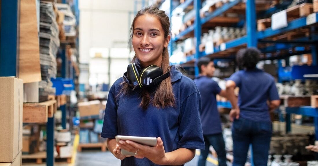 Woman standing in warehouse, smiling and holding a pad of paper in hand.