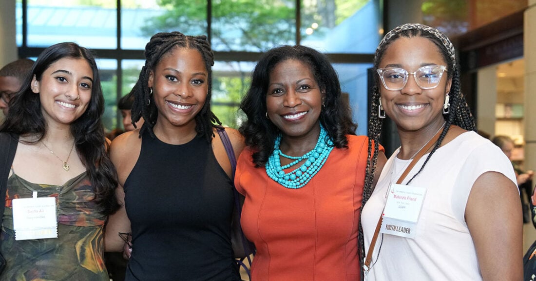 Four women standing together and smiling at a gathering. The woman in the orange dress with a turquoise necklace is centered in the group. Two women stand on her left and one woman on her right.