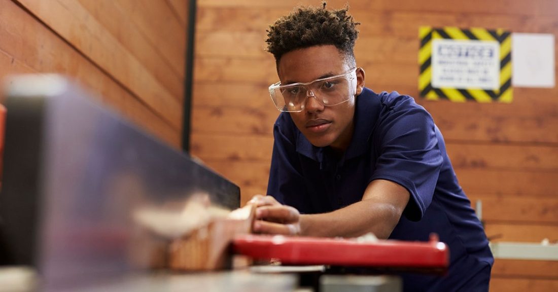 Young Black man wearing safety glasses works in a woodshop, learning over a saw with a piece of wood in hand.