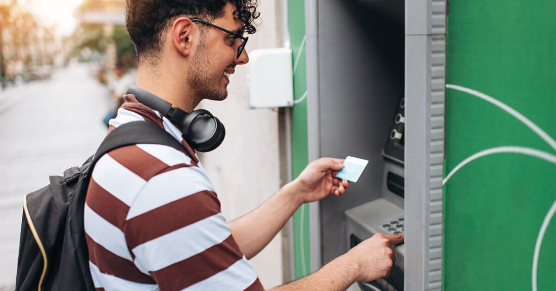 A young man with a black backpack and headphones around his neck stands in front of an ATM. He's smiling and is holding his banking card in front of him, ready to use it at the ATM.