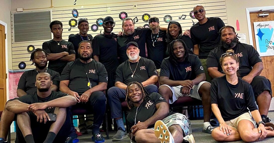 Fifteen people, all in matching black T-shirts, pose for a photo in a classroom-like setting. Some are peope of color, a few are white. Most are men, a few are women. Some individuals are smiling, others are not.