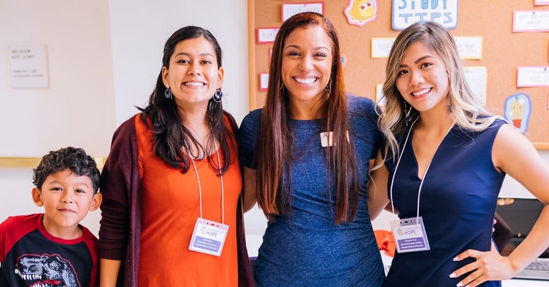 Three women and one young boy standing in a room, arm in arm, wearing nametags and smiles.