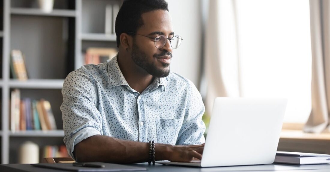 Black man with glasses sits inside at a desk, looking at his laptop screen.