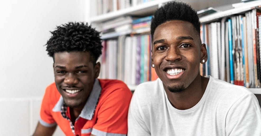 Two young black men sit in front of a bookcase, smiling at the camera.