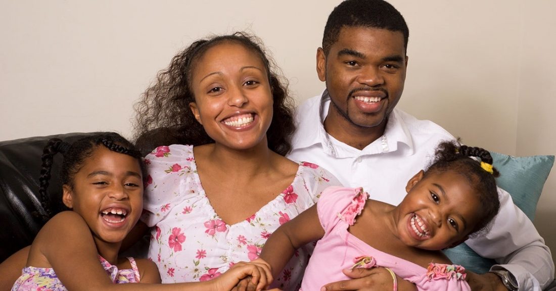 A Black family of four, including two young daughters, sitting on a couch and smiling.
