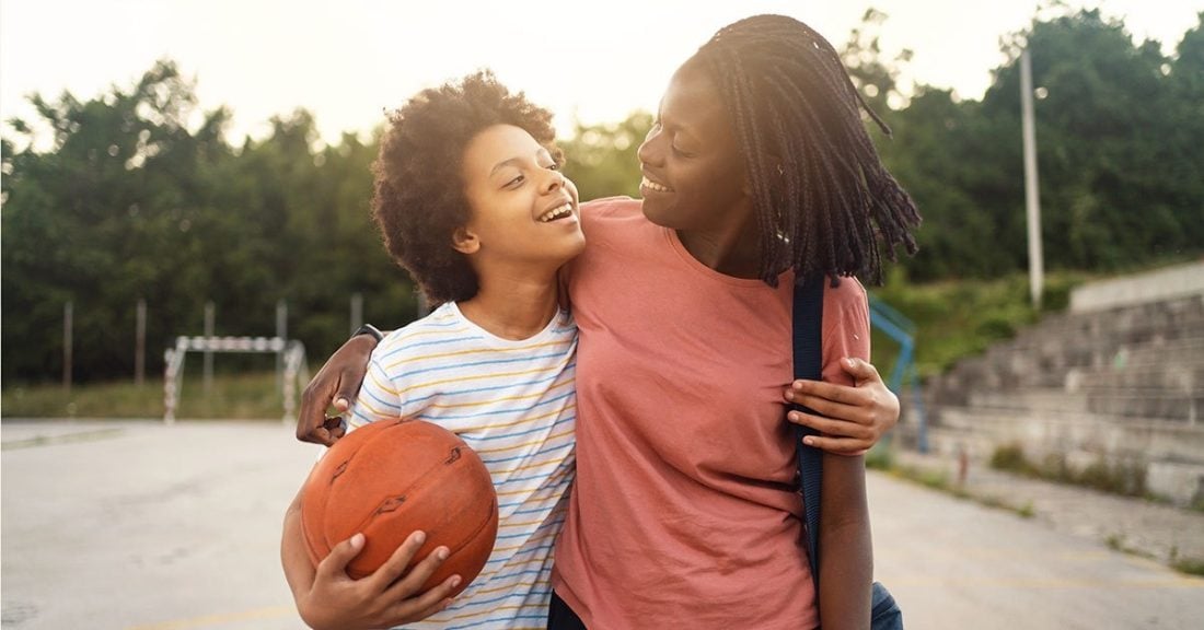 A mother and her child are standing outdoors, each with one arm wrapped around the other. They are looking at each other and smiling. The child has a basketball in hand.