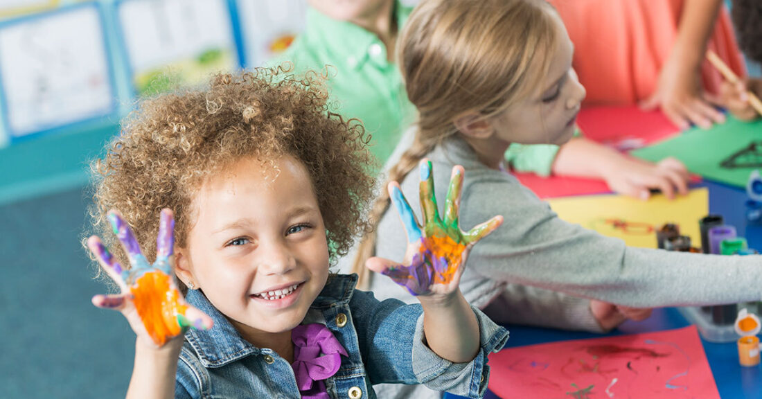 A little girl during an art activity at a child care center. She is sitting at a table with other children painting pictures. She is looking at the camera, smiling, holding up her hands covered in paint.