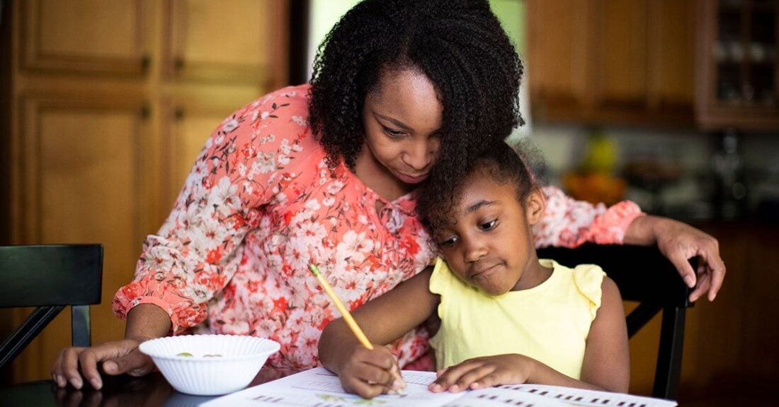 An adult woman sits in a kitchen with a young girl who is writing in an open notebook.