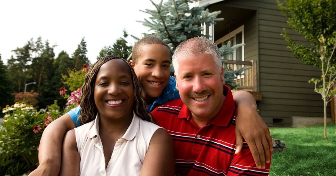 Family of three, two adults and one child, sit outdoors, smiling, in front of a house.