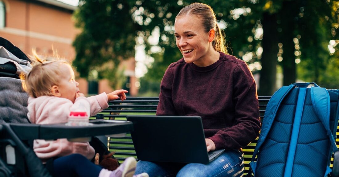 Woman works on a laptop while a small toddler sits in a stroller nearby.