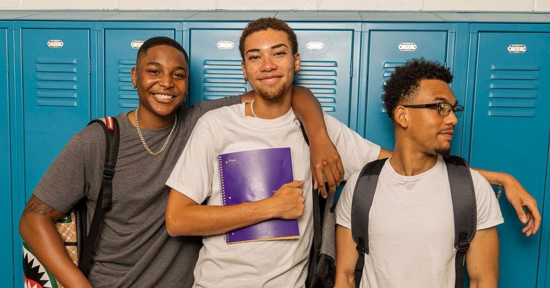 Three young men of color, wearing bookbags, stand against blue lockers