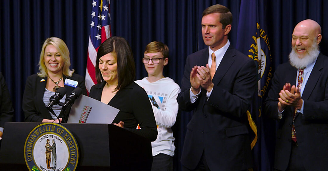 Five happy people, all white, standing in front of blue pipe and drape, clapping. One woman stands at a podium, smiling, with a "Kentucky Voices for Health" folder open before her.