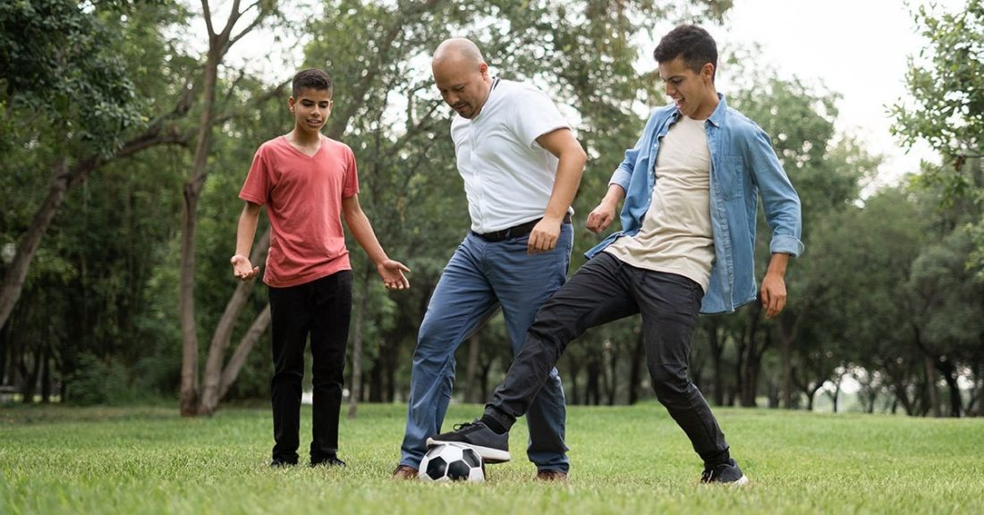 A dad plays soccer with his two young boys; all three are outdoors.