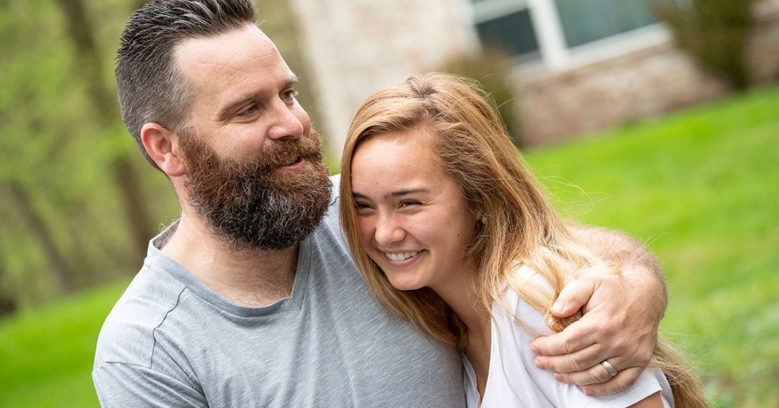 White father with beard has arm around teen daughter with long blonde hair. They are outdoors and wearing tee-shirts.