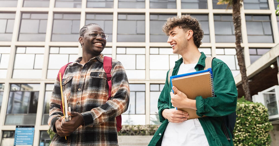 Two young men are standing outside their college or university with notebooks in hand, smiling at one another.