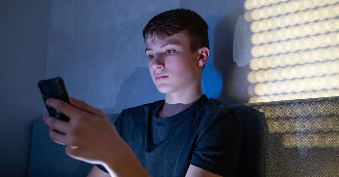 A teenage boy sits on a couch in a dimly lit room, illuminated by the light from his smartphone as he looks at the screen.