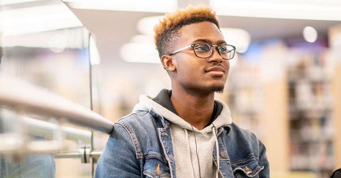 A Black, male teenager smiles, wearing glasses and a denim jacket.