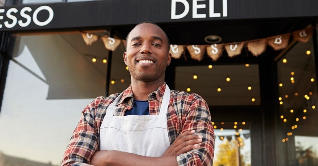Black man wearing a white apron stands outside a food shop with his arms crossed across his chest.
