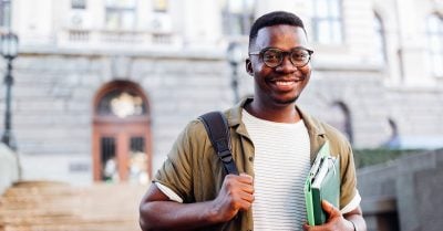 一个年轻的黑人对着镜头微笑。他戴着眼镜，穿着短袖衬衫。他一个肩膀上挎着一个背包，另一只胳膊上抓着一本书和一个文件夹。