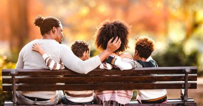 The image is a photo of a Black family of four — taken from behind — sitting  on an outdoor bench, with their arms around each other. The father, on the far left, has dreadlocks pulled into a bun, while the mother has textured, natural hair. They are joined by their two small sons.