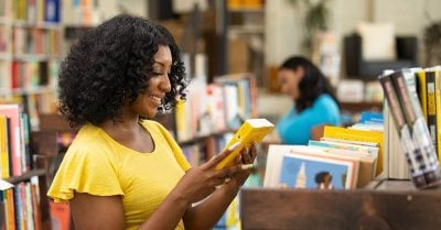 A young Black woman, wearing a yellow short-sleeved shirt, smiles while examining a book in a library.