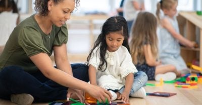 A young female childcare worker of color sits with a pre-school-age Brown girl in a day care setting. The two are engaged in a learning activity.