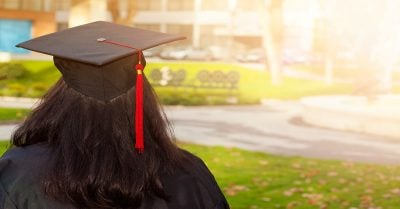 一位戴着毕业帽的年轻黑人女性背对着镜头站着。她戴着毕业帽，穿着毕业礼服，望向阳光明媚的四合院。