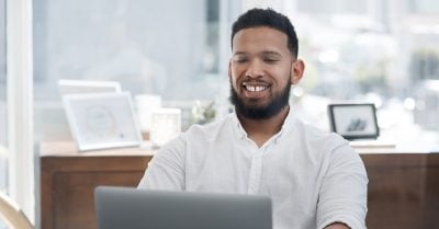 Man of color with beard and white shirt sits at a laptop in an office.