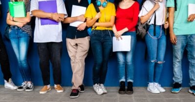 A diverse group of high school students, shot from the neck down, stand lined up against lockers in a school hallway.