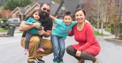 A young Indigenous family of four — a father, mother, daughter and son — crouch down on a residential street for a family photo.