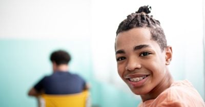 A young, Black teenage boy smiles into the camera, while sitting in a classroom setting. The boy wears braces and his hair is styled in braids.