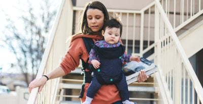 A young Latino mother walks down a flight of outdoor steps; her infant child is nestled in front of her, strapped into a baby-wearing device.