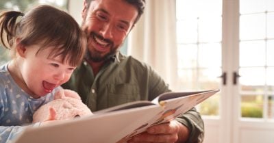 A young girl smiles as her father, also smiling, holds open a picture book for her to explore.