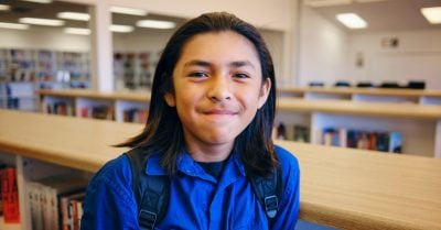 A Native American youth wears a backpack and smiles at the camera; he's in a library