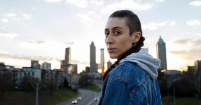 A young person of Hispanic descent — wearing a hoodie with a jean jacket — looks over their shoulder into the camera. They are standing outside, and the Atlanta skyline is in the background.