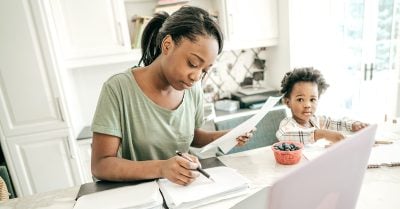 Black mother sits at her kitchen counter with a pen and paper in had; her young child sits next to her coloring