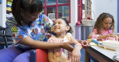 A black woman wearing medical scrubs smiles next to two young girls of color who are playing at a table.