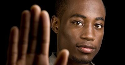 A young Black male gazes into the camera. His open hand is raised in front of him, in a gesture likely to mean “stop.”