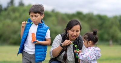 A young Native mother plays outside with her two small children, showing them how to blow bubbles with a bubble wand and solution.
