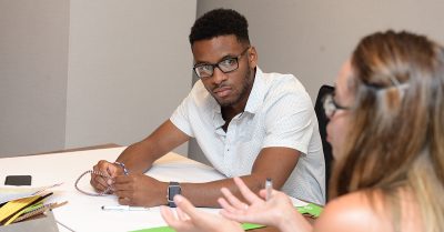 A young black man looks at a woman as she speaks at a meeting