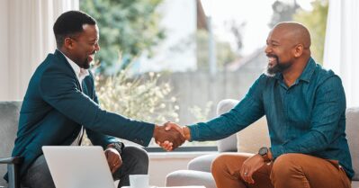 Two Black men shake hands in a professional setting, while smiling at each other.