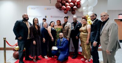 The image depicts s diverse group of Black and Brown young men and women on the red carpet of a formal event. The group smiles while standing in front of a banner displaying the logo for the Community Violence Intervention Leadership Academy.