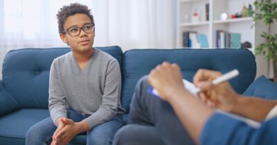 The scene shows a young boy of color, wearing glasses, sitting on a blue sofa. He appears to be speaking with an adult counselor — whose face is not shown — who holds a clipboard and pen in their lap.