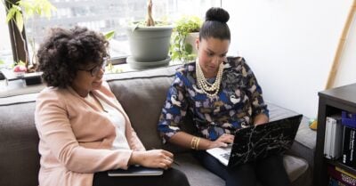 Two young Black women — dressed professionally — sit side-by-side on a sofa in an office setting. They peer down at a laptop, while one holds a notebook and pen.