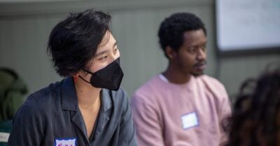 In the foreground, a young Asian woman wearing a mask and a nametag listens intently in a lecture setting. A young Black man sits behind her, also listening intently.