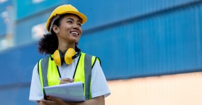 A young Black woman wears a yellow safety vest, yellow earphones/ear protectors and a yellow hardhat. She smiles as she holds a clipboard in front a blue factory wall.
