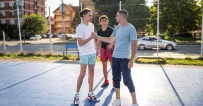 Three adolescent males gather on an outdoor basketball court. Two of them exchange a friendly handshake grip, while the third looks on.