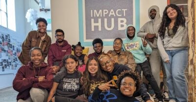 A group of Black and Brown young people gather for a photo in a casual, indoor setting. Behind them is a sign that reads, “Impact Hub.”