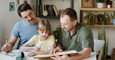 Two white dads site on either side of their daughter. A book open and pencils in hand, they help her study at their home.