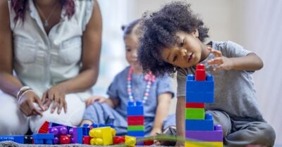 A play scene, with a toddler playing with colorful plastic blocks with another toddler and a woman sitting behind.